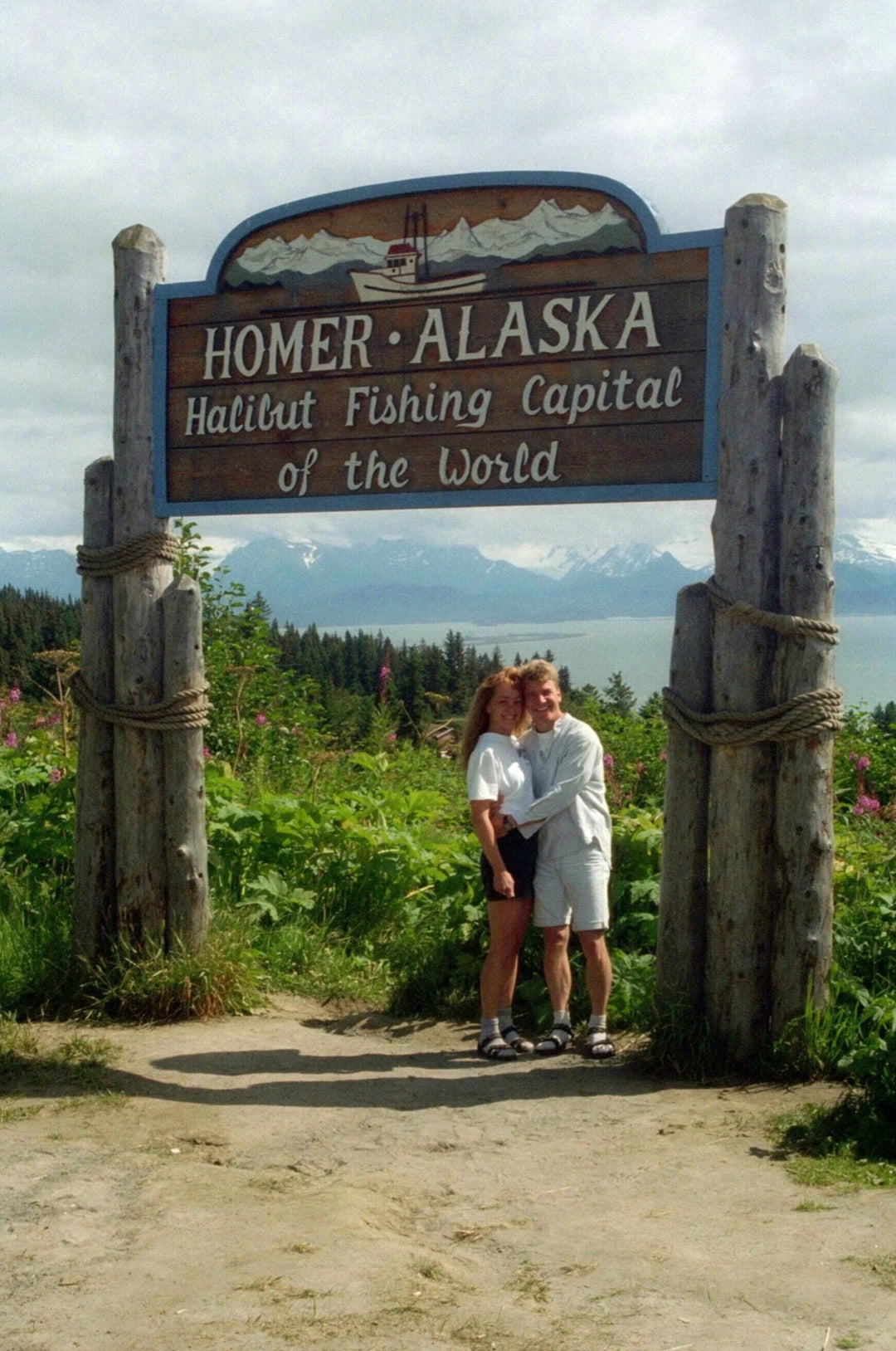 Two people standing in front of a sign for Homer, Alaska, with mountains in the background.