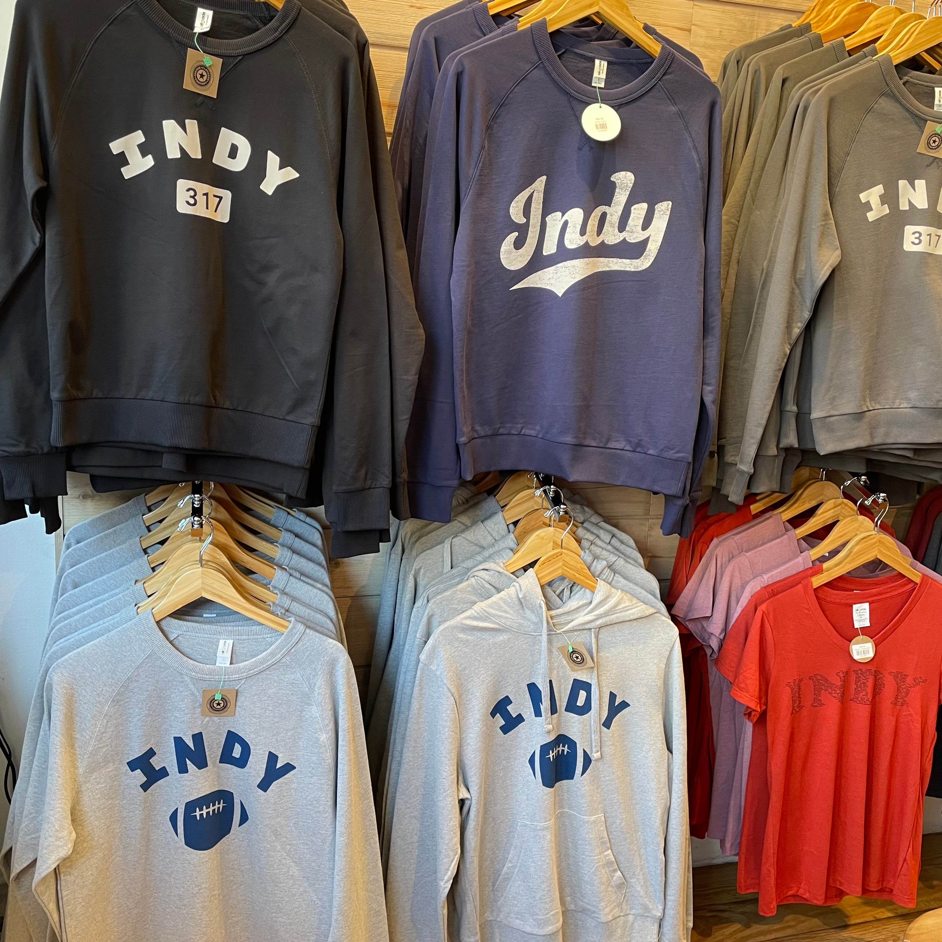 Collection of sweatshirts and t-shirts with 'INDY' branding on hangers against a wooden wall.