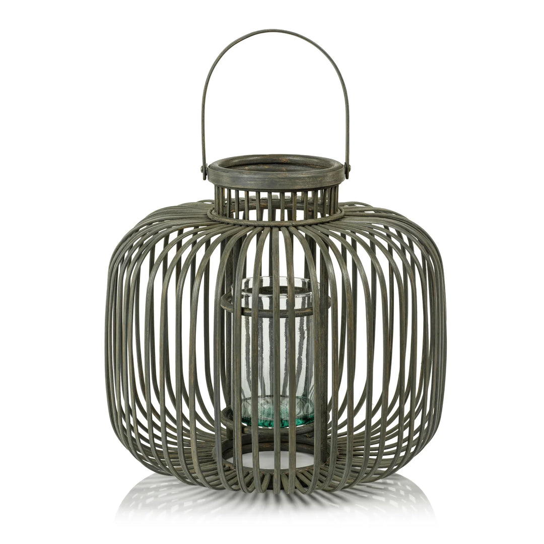 A rustic metal lantern with a wire cage design, a top handle, and a clear glass candle holder inside, shown on a white background with a soft reflection | Artisans